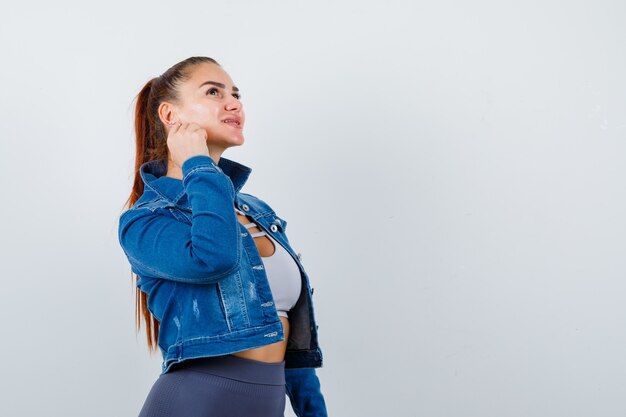 Giovane donna in alto, giacca di jeans con pugno sulla guancia e sguardo pensieroso, vista frontale.