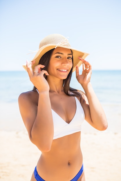 Giovane donna in bikini che indossa il cappello di paglia bianco godendo le vacanze estive in spiaggia. Ritratto di bella donna latina rilassante in spiaggia con occhiali da sole.