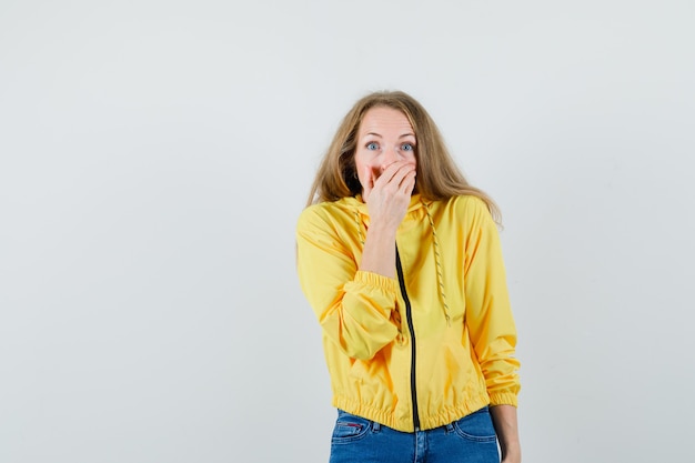 Giovane donna in bomber giallo e jeans blu che copre la bocca con la mano e guardando sorpreso, vista frontale.