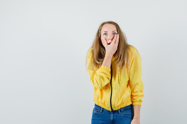 Giovane donna in bomber giallo e jeans blu che copre la bocca e che sembra sorpresa, vista frontale.