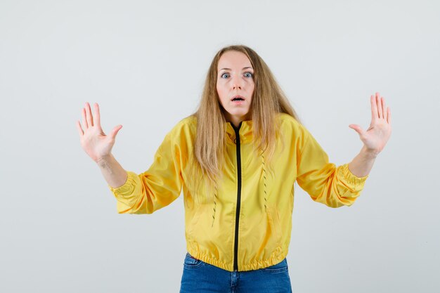 Giovane donna in bomber giallo e jeans blu che mostra il gesto di resa e che sembra spaventata, vista frontale.