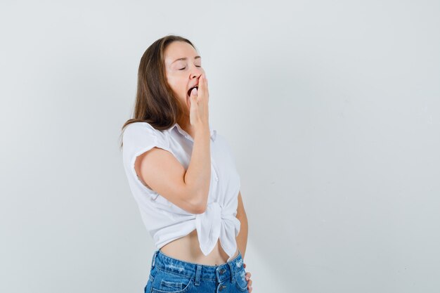 Giovane donna in camicetta bianca che sbadiglia con la mano sulla bocca e sembra assonnata, vista frontale. spazio per il testo