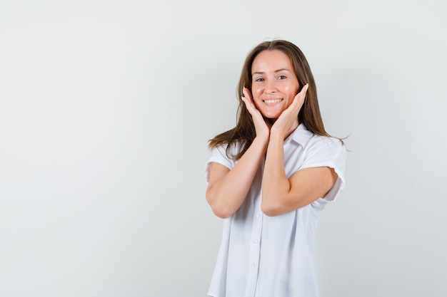 Giovane donna in camicetta bianca tenendo le mani sul viso e guardando felice