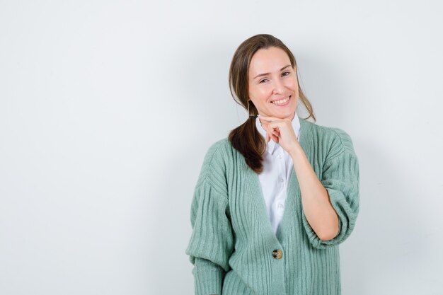 Giovane donna in camicetta, cardigan che sostiene il mento a portata di mano e sembra felice, vista frontale.