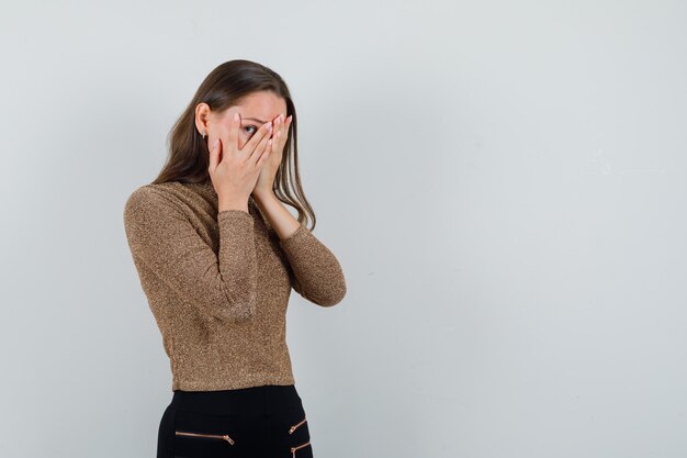 Giovane donna in camicetta dorata che guarda attraverso le dita e che sembra spaventata