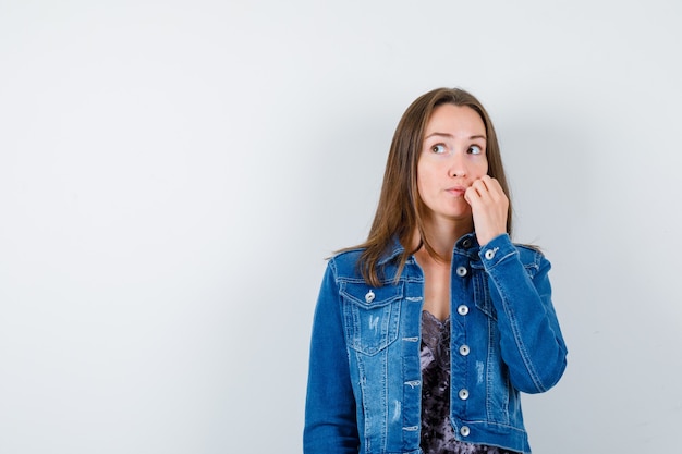 Giovane donna in camicetta, giacca di jeans che graffia la guancia mentre distoglie lo sguardo e sembra pensierosa, vista frontale.