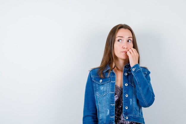 Giovane donna in camicetta, giacca di jeans che graffia la guancia mentre distoglie lo sguardo e sembra pensierosa, vista frontale.