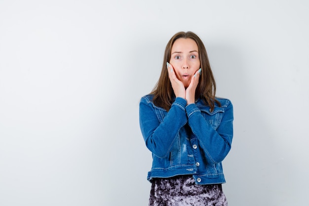 Giovane donna in camicetta, giacca di jeans con le mani sulle guance e sguardo perplesso, vista frontale.