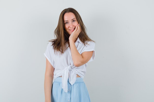 Giovane donna in camicetta, gonna in posa con la mano sulla guancia e dall'aspetto carino