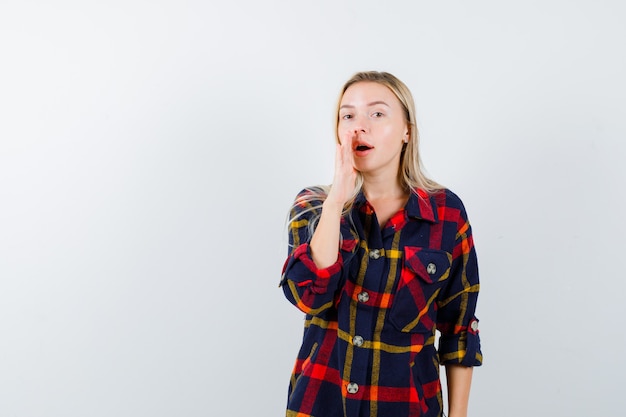 Giovane donna in camicia a quadri che dice il segreto con la mano e che sembra seria, vista frontale.