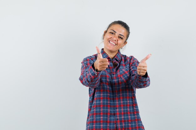 Giovane donna in camicia a quadri che mostra i doppi pollici in su e che sembra felice, vista frontale.