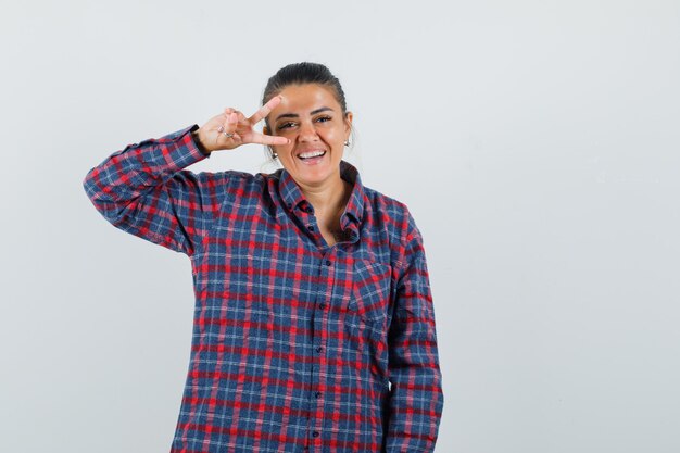 Giovane donna in camicia a quadri che mostra il segno di v sull'occhio e che sembra felice, vista frontale.