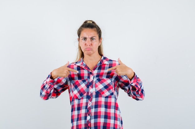 Giovane donna in camicia a quadri che punta a se stessa e che sembra seria, vista frontale.