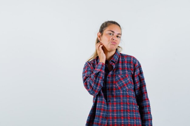 Giovane donna in camicia a quadri in posa mentre esamina la sua pelle e sembra allettante, vista frontale.