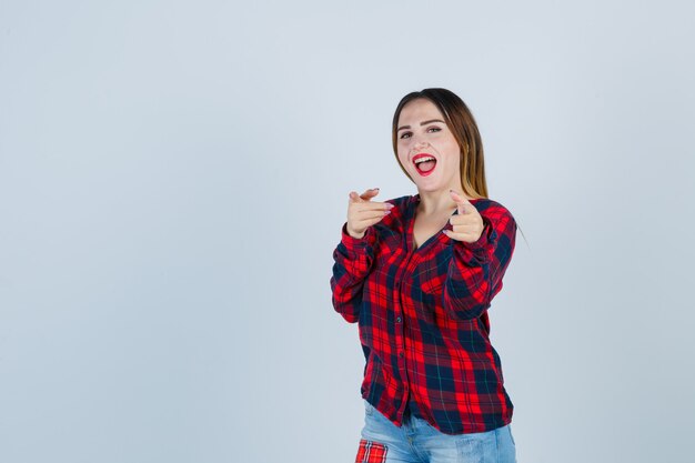 Giovane donna in camicia a quadri, jeans che puntano sul davanti, bocca aperta e aspetto delizioso, vista frontale.