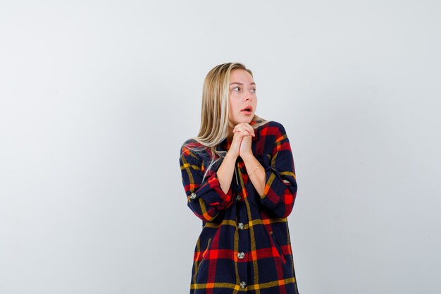 Giovane donna in camicia a quadri tenendo le mani giunte sul petto mentre guarda lontano e sembra spaventata, vista frontale.