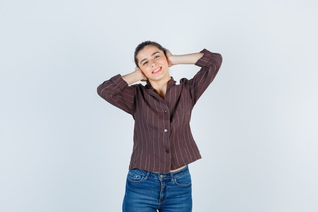 Giovane donna in camicia a righe, jeans premendo le mani sulle orecchie e guardando allegra, vista frontale.