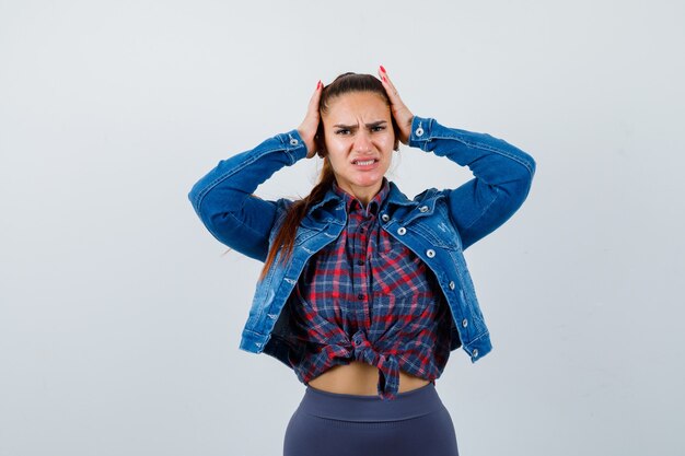 Giovane donna in camicia a scacchi, giacca di jeans con le mani sulla testa e sguardo doloroso, vista frontale.