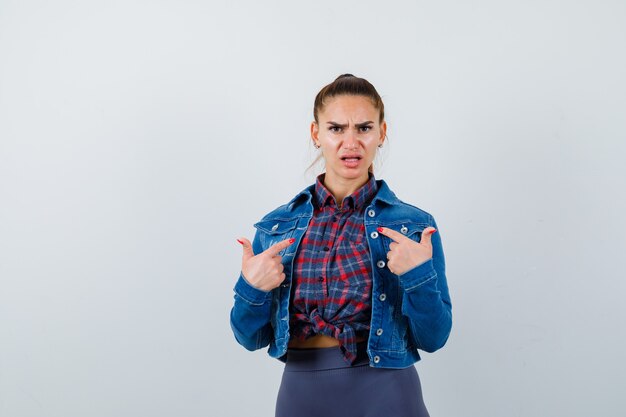 Giovane donna in camicia a scacchi, giacca, pantaloni che punta a se stessa e sembra impotente, vista frontale.