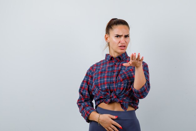 Giovane donna in camicia a scacchi, pantaloni che gesticolano con il palmo verso l'esterno per fermarsi e sembrano seri, vista frontale.