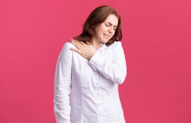 Giovane donna in camicia bianca alla ricerca di malessere toccando la sua spalla sensazione di dolore in piedi sopra il muro rosa