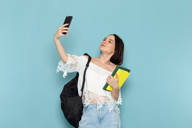 giovane donna in camicia bianca blue jeans e borsa nera in possesso di quaderni prendendo un selfie sul blu