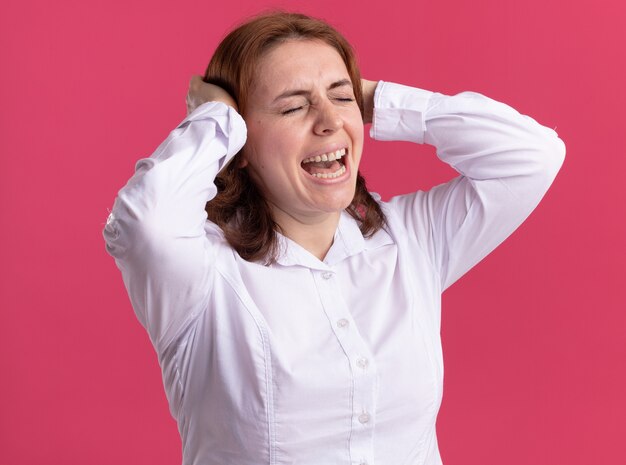Giovane donna in camicia bianca che grida con espressione infastidita con le mani sulla sua testa in piedi sopra il muro rosa