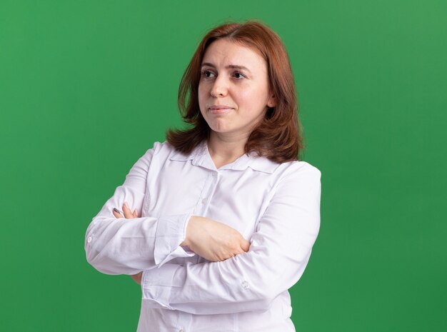 Giovane donna in camicia bianca che osserva da parte con la faccia seria con le braccia incrociate in piedi sopra la parete verde