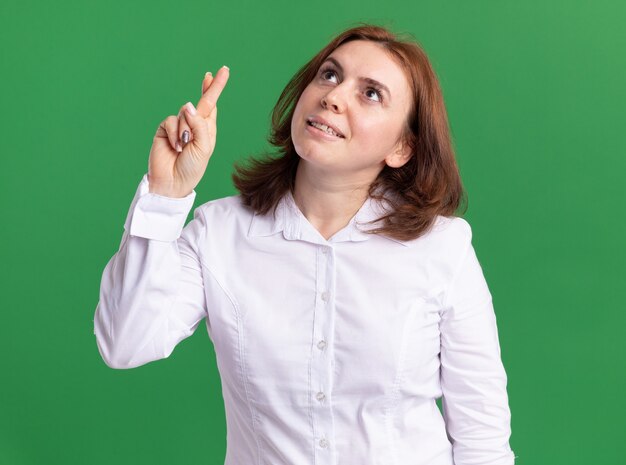 Giovane donna in camicia bianca che osserva in su con il sorriso sulle dita incrociate del viso in piedi sopra la parete verde