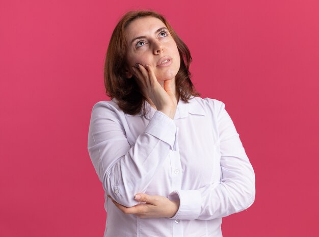 Giovane donna in camicia bianca che osserva in su con la mano sulla sua guancia pensando in piedi sopra il muro rosa