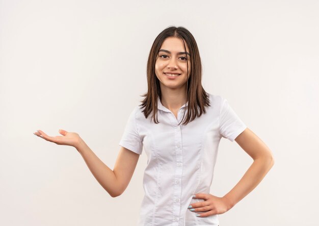 Giovane donna in camicia bianca che presenta qualcosa con il braccio della sua mano sorridendo allegramente in piedi sopra il muro bianco