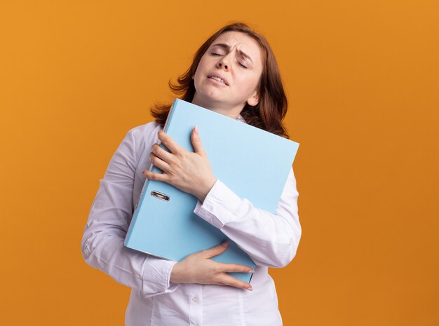 Giovane donna in camicia bianca che tiene cartella cercando stanco e sovraccarico di lavoro in piedi sopra la parete arancione