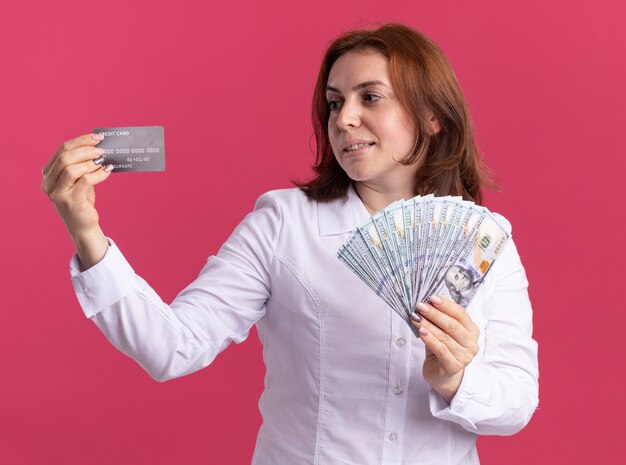 Giovane donna in camicia bianca che tiene contanti guardando la carta di credito in mano sorridente felice e positivo in piedi oltre il muro rosa