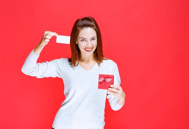 Giovane donna in camicia bianca che tiene in mano una piccola scatola regalo rossa e presenta il suo biglietto da visita