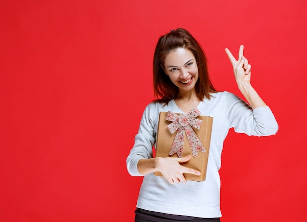 Giovane donna in camicia bianca che tiene in mano una scatola regalo di cartone e mostra un segno positivo con la mano