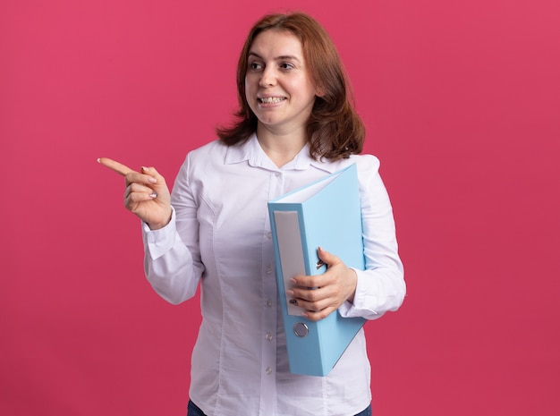 Giovane donna in camicia bianca che tiene la cartella guardando da parte che punta con il dito indice a lato sorridente allegramente in piedi sopra il muro rosa