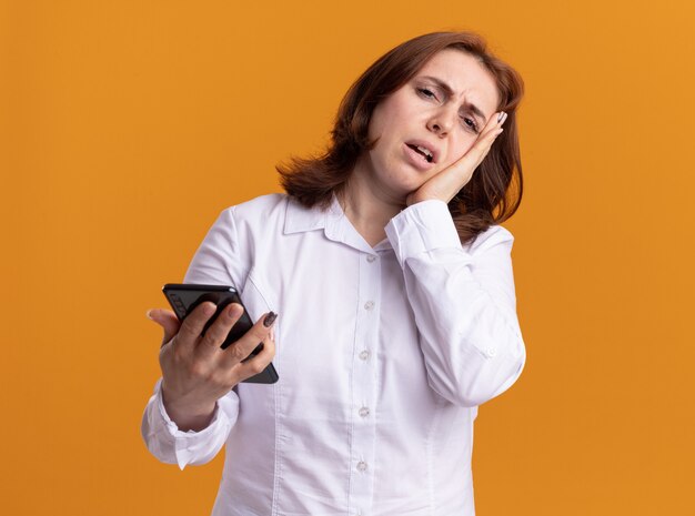 Giovane donna in camicia bianca con lo smartphone guardando davanti confuso e scontento in piedi sopra la parete arancione