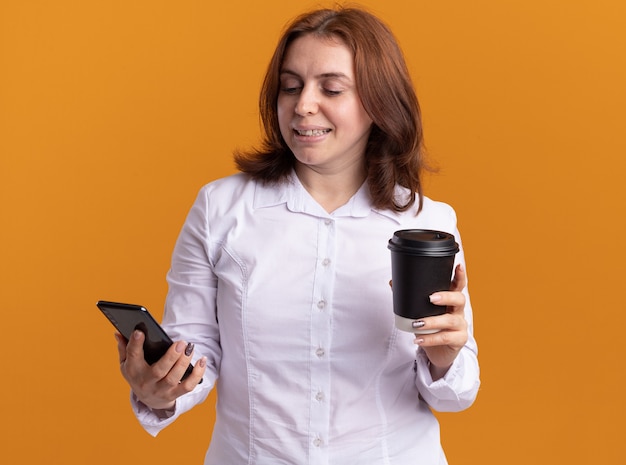 Giovane donna in camicia bianca con lo smartphone tenendo la tazza di caffè guardando il suo cellulare con il sorriso sul viso in piedi sopra la parete arancione
