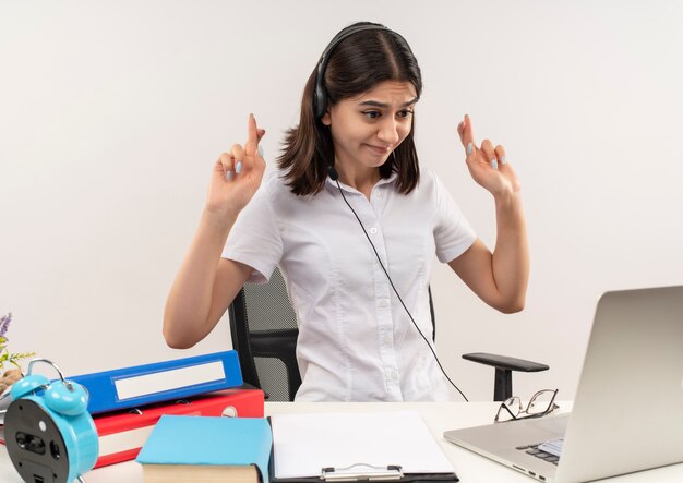 Giovane donna in camicia bianca e cuffie con un microfono seduto al tavolo con cartelle guardando lo schermo del suo laptop in videochiamata rendendo desiderabile desiderio incrociando le dita sul muro bianco