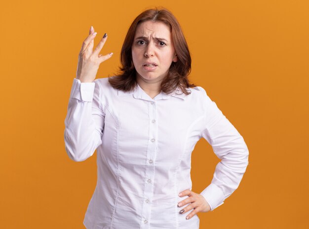 Giovane donna in camicia bianca guardando davanti essendo scontento alzando la mano per il dispiacere in piedi oltre il muro arancione