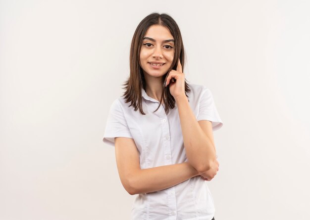 Giovane donna in camicia bianca guardando in avanti sorridente fiducioso in piedi oltre il muro bianco