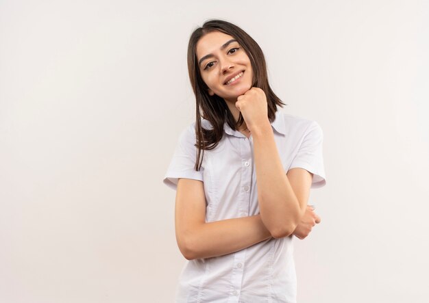 Giovane donna in camicia bianca guardando in avanti sorridente fiducioso in piedi oltre il muro bianco