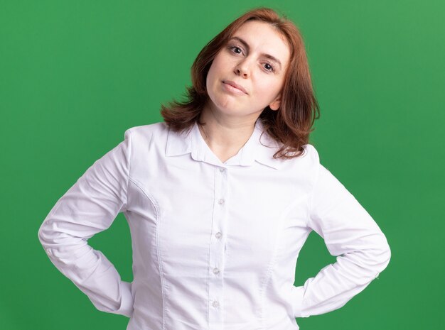 Giovane donna in camicia bianca guardando la parte anteriore con seria espressione fiduciosa in piedi sopra la parete verde