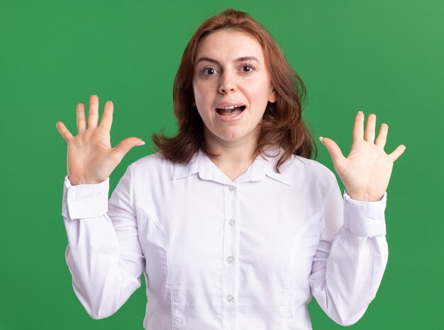 Giovane donna in camicia bianca guardando la parte anteriore sorridente con la faccia felice alzando le mani in piedi sopra la parete verde