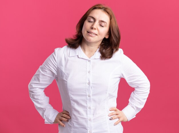 Giovane donna in camicia bianca sorridente con la faccia felice con gli occhi chiusi in piedi sul muro rosa