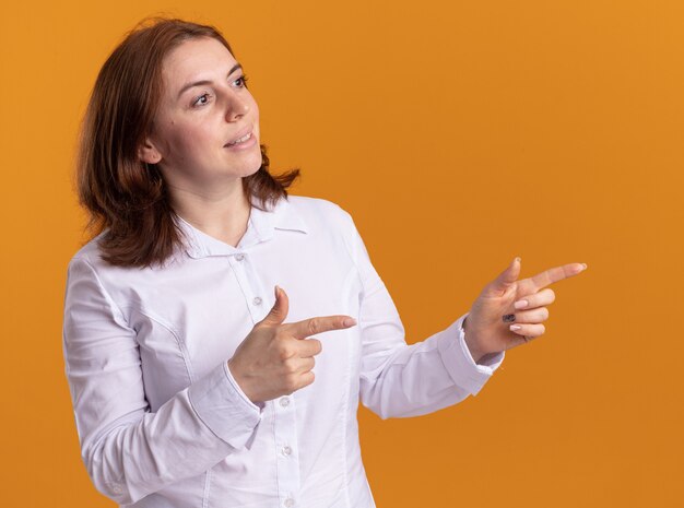 Giovane donna in camicia bianca sorridente fiducioso guardando da parte che punta con il dito indice a lato in piedi sopra la parete arancione