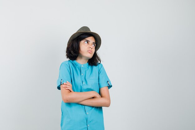 Giovane donna in camicia blu, cappello in piedi con le braccia incrociate mentre guarda da parte e sembra complicata