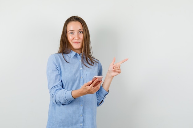 Giovane donna in camicia blu che tiene il telefono cellulare mentre punta lontano e sembra curioso