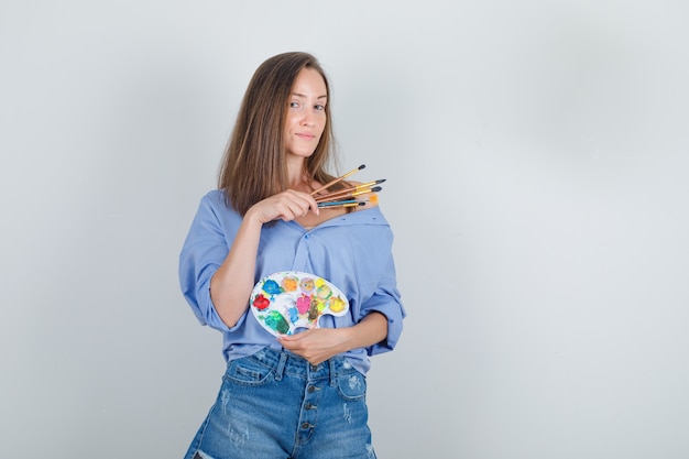 Giovane donna in camicia blu, pantaloncini tenendo gli strumenti di pittura e guardando orgoglioso