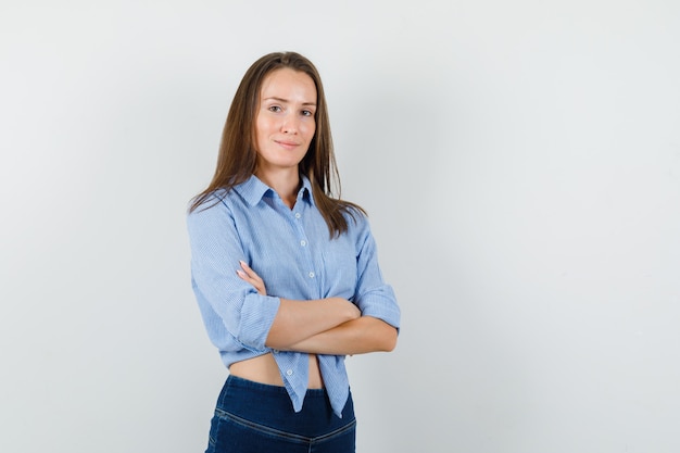 Giovane donna in camicia blu, pantaloni in piedi con le braccia incrociate e che sembra allegra.